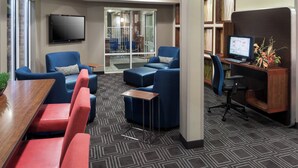 A modern hotel lobby with a large wooden table, red and blue seating, a flat-screen TV, a computer desk, and a patterned carpet. - TownePlace Suites by Marriott Little Rock West (Little Rock)