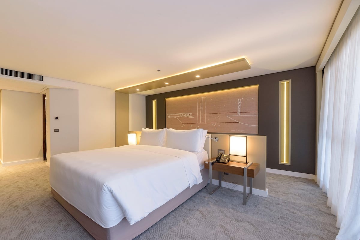 A modern hotel room with a large bed, a bedside table with a lamp, a wall-mounted artwork, and a door.