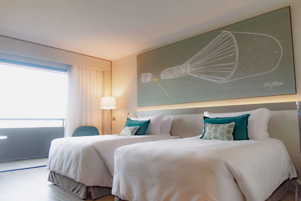 A hotel room with two beds, a large abstract painting on the wall, a window with curtains, and a floor lamp.