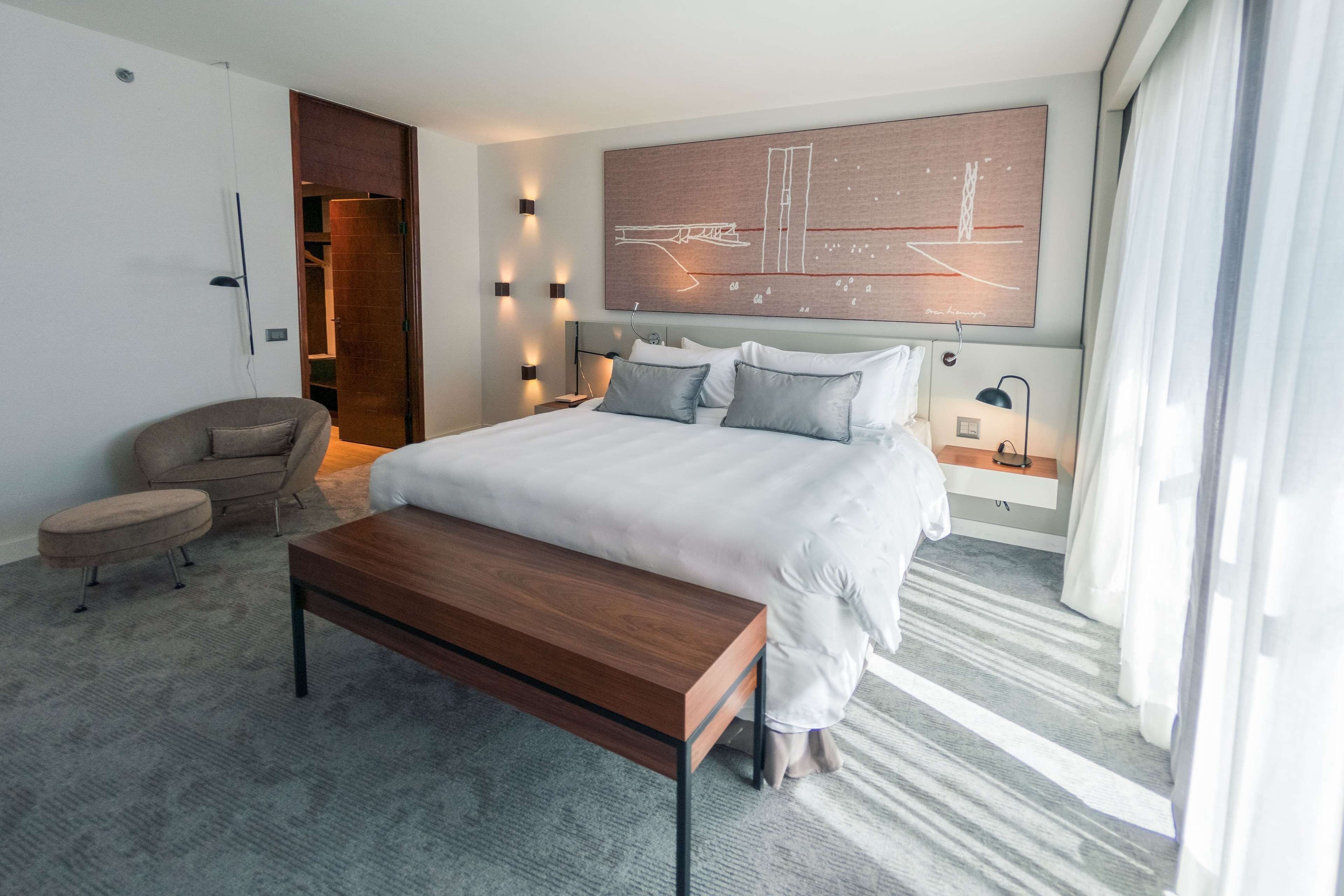 Una habitación de hotel moderna con una cama grande, un banco de madera, una silla y un cuadro abstracto de gran tamaño en la pared.