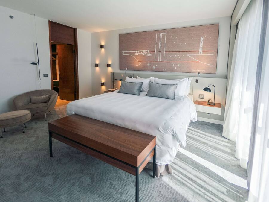 Una habitación de hotel moderna con una cama grande, un banco de madera, una silla y un cuadro abstracto de gran tamaño en la pared.