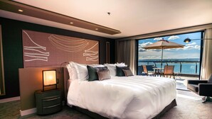 Una habitación de hotel con una cama grande, una mesita de noche, vistas al mar y un balcón con sillas y una sombrilla.
