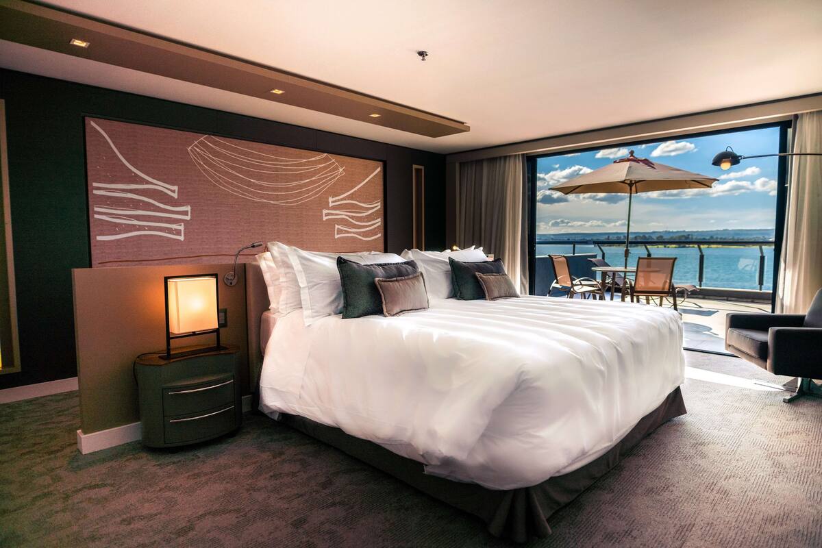 A hotel room with a large bed, a nightstand, a view of the sea, and a balcony with chairs and an umbrella.