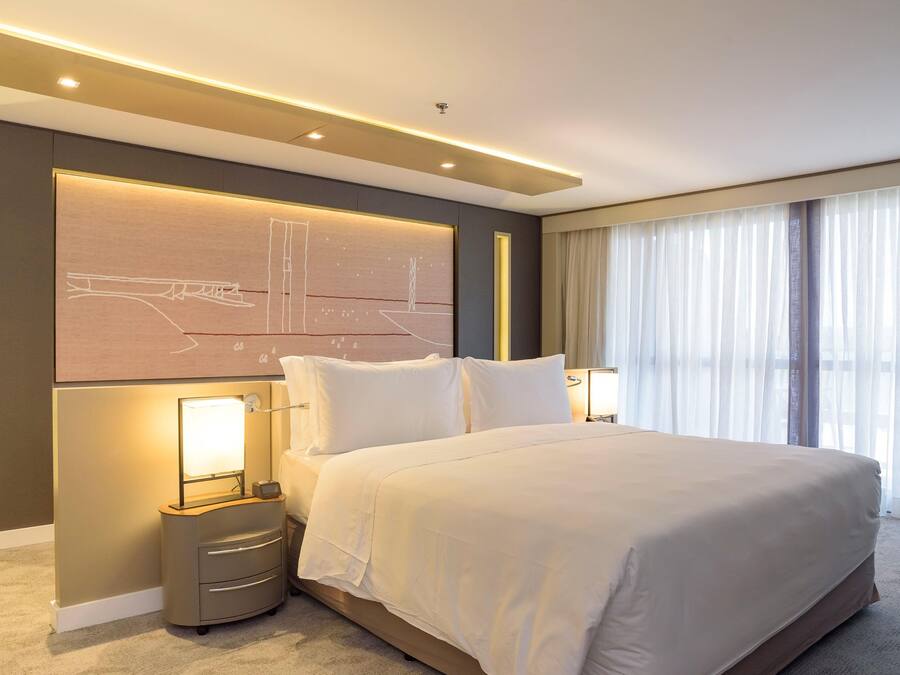 Quarto de hotel moderno com uma cama grande, abajures, cabeceira com design de paisagem urbana e uma janela ampla com cortinas leves.