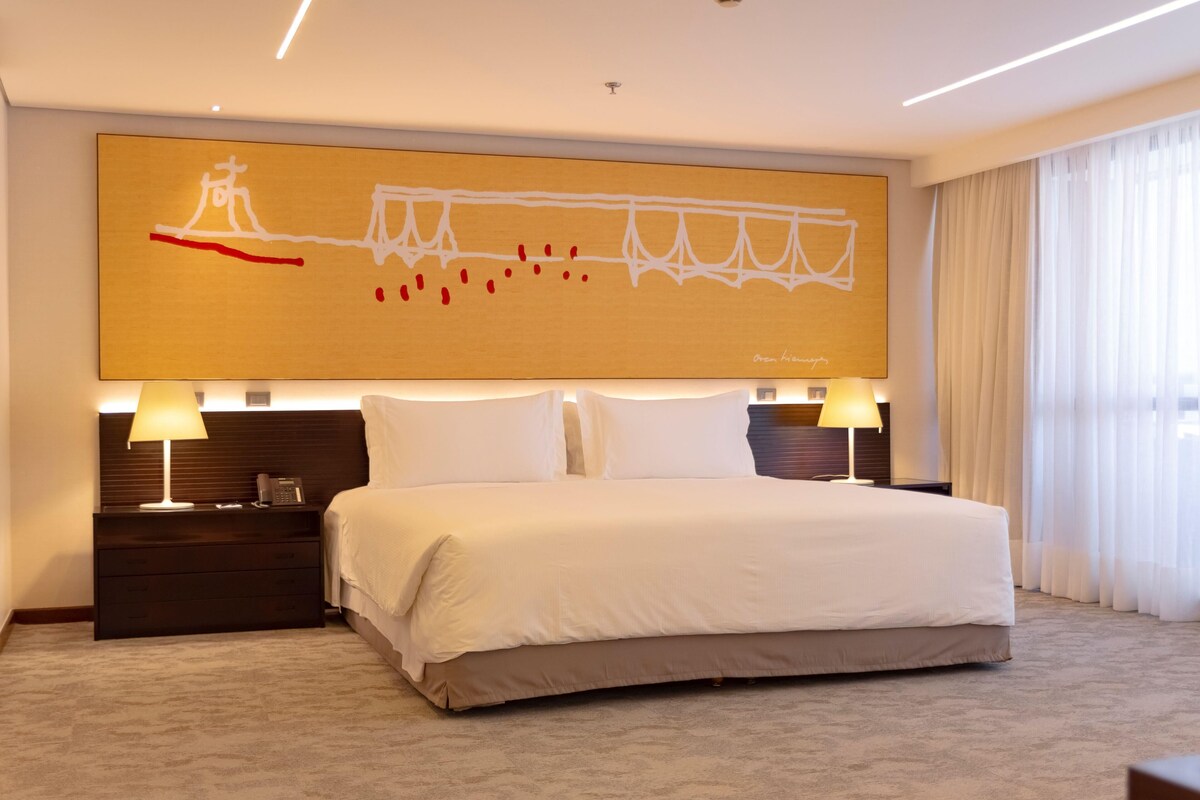 A modern hotel room with a large bed, two bedside tables with lamps, a large abstract painting on the wall, and a view of the city through sheer curtains.