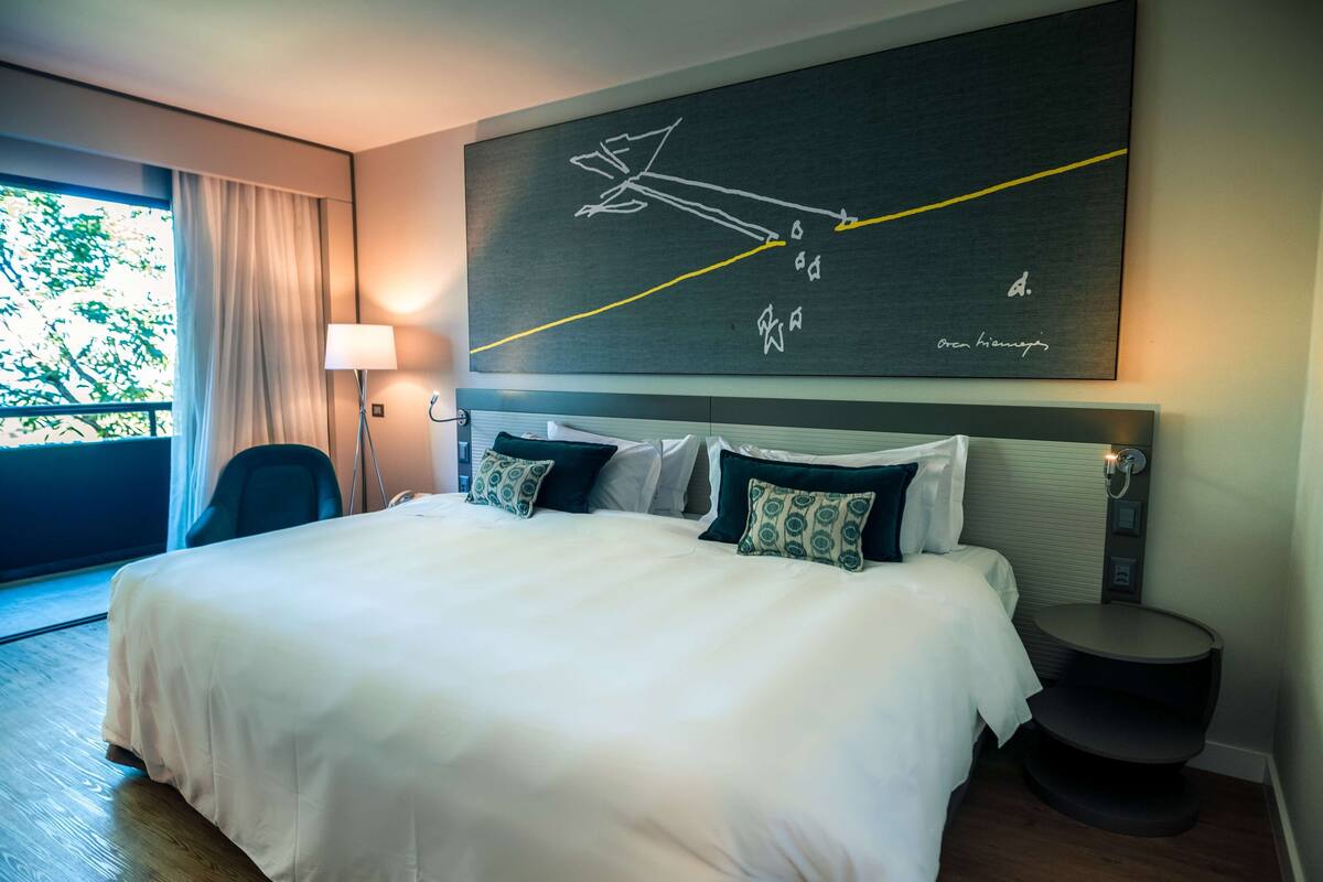 A hotel room with a large bed, a headboard with a clock, a bedside table, and a view of greenery outside.