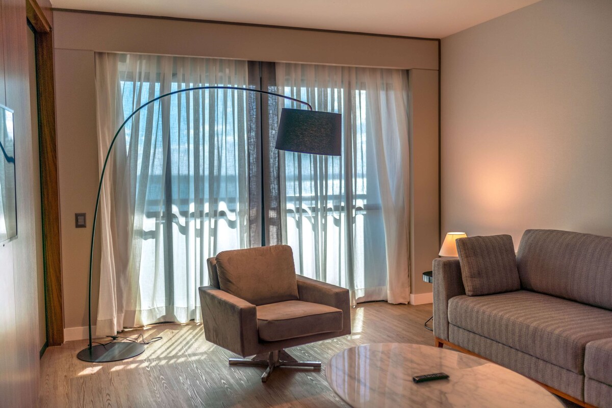 A modern living room with a sofa, armchair, and floor lamp. A TV is mounted on the wall, and a balcony with curtains is visible through the window.