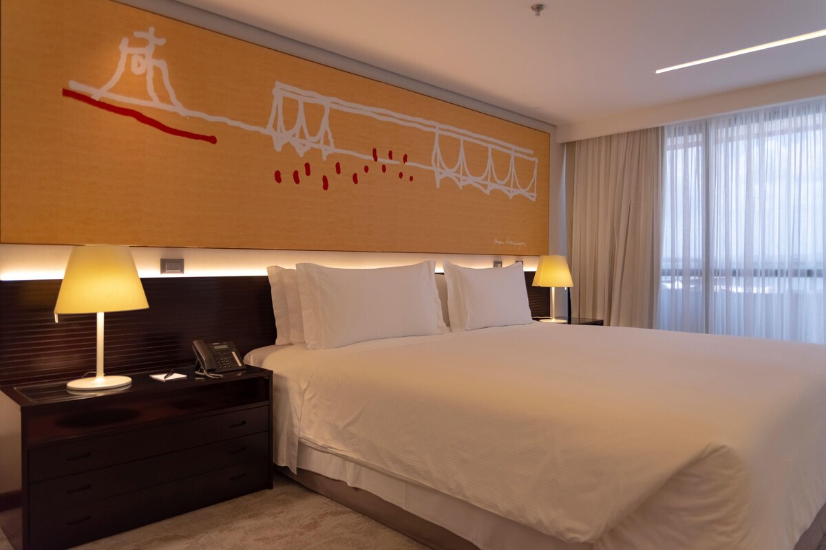 A hotel room with a large bed, two bedside tables with lamps, a wall-mounted artwork, and a large window with curtains.