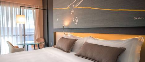 Habitación de hotel con una cama grande, dos almohadas, una mesita de vidrio, una silla y una pared decorada con arte abstracto.