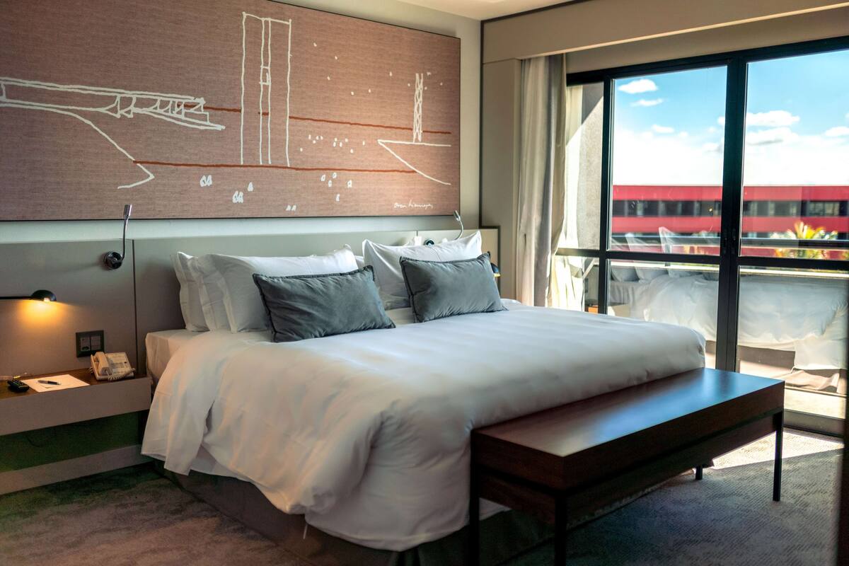 A modern hotel room with a large bed, bedside table, and a view of the city through large windows.