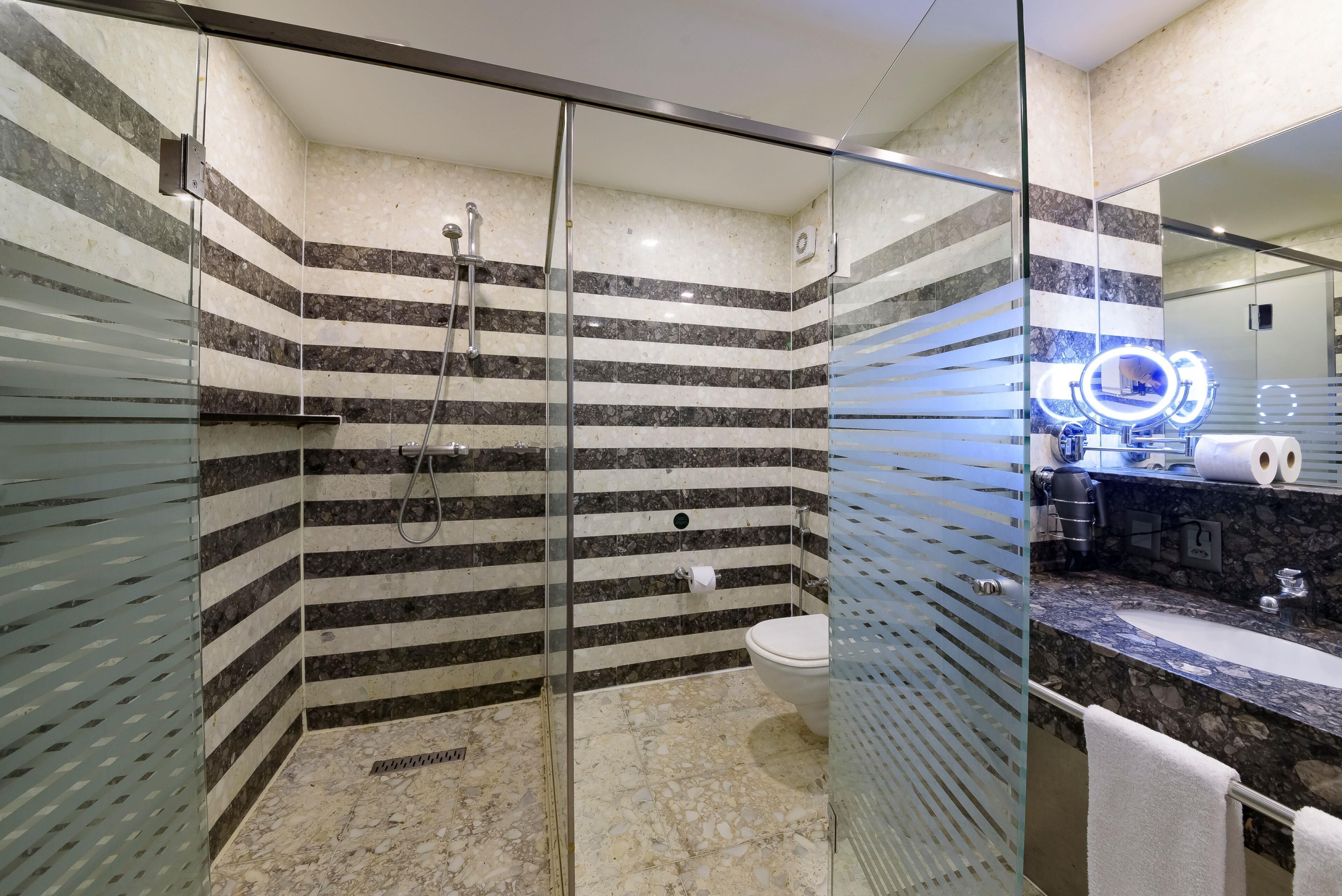 Un baño moderno con ducha de vidrio, piso de mármol y tocador con lavamanos y espejo.