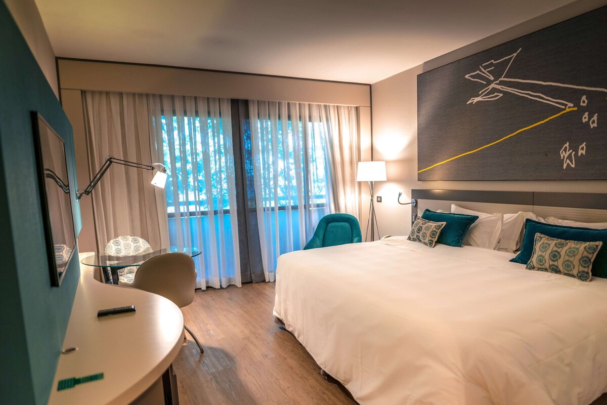A hotel room with a large bed, a desk, a chair, a window with curtains, and a wall art piece.
