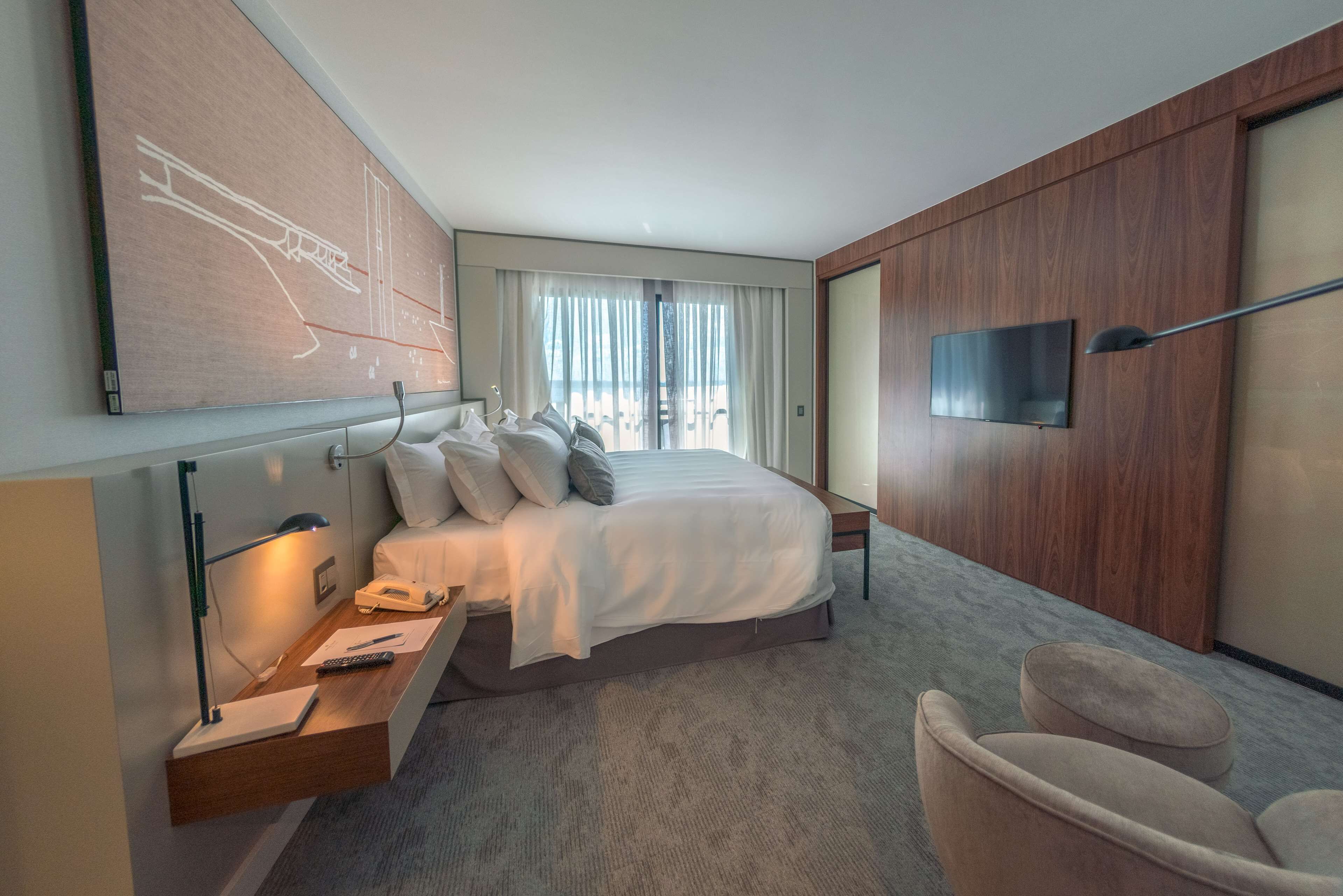 Ein modernes Hotelzimmer mit einem großen Bett, einem Schreibtisch mit Lampe, einem Flachbildfernseher und einem großen Fenster mit Vorhängen.