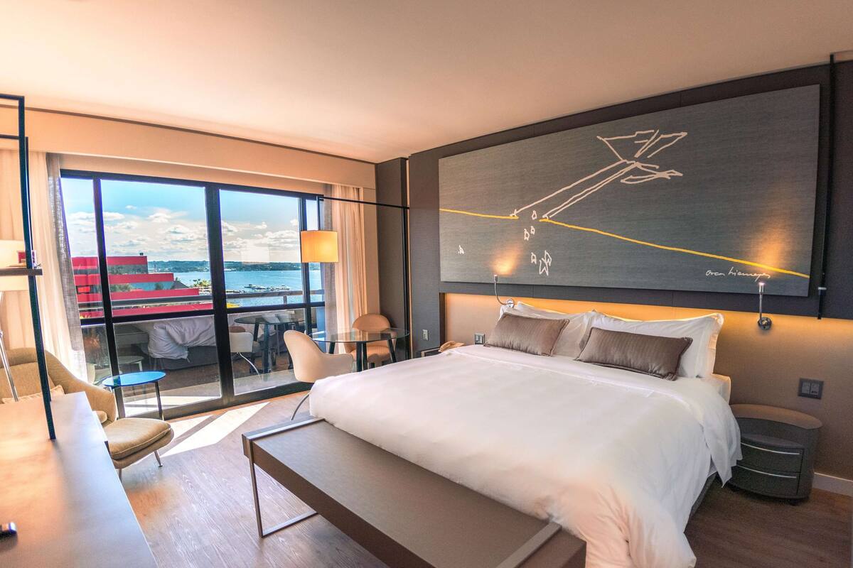 A modern hotel room with a large bed, a desk, and a balcony with a view of the sea.