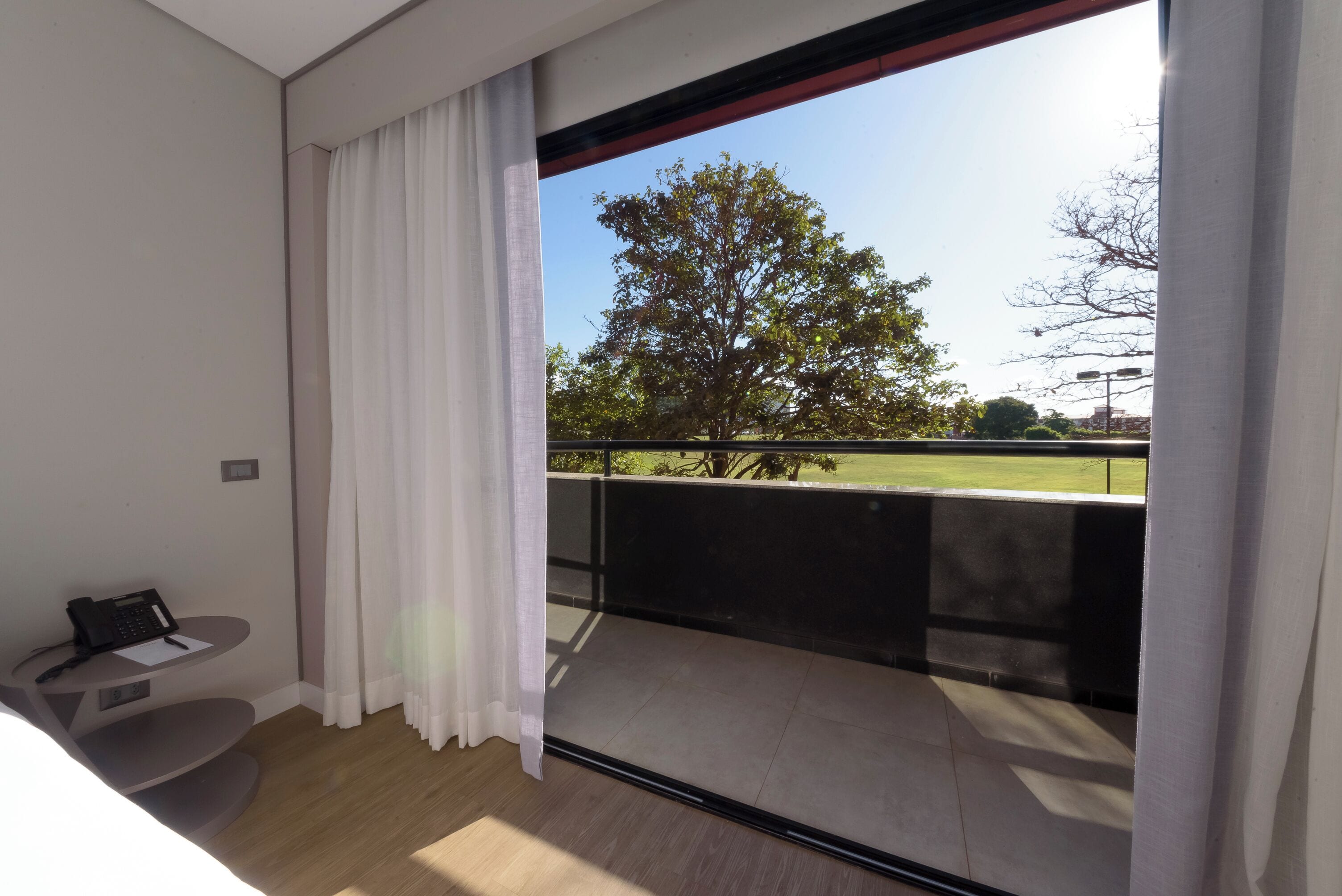 Habitación de hotel moderna con un ventanal grande, un escritorio pequeño con teléfono y vista a un parque y árboles.