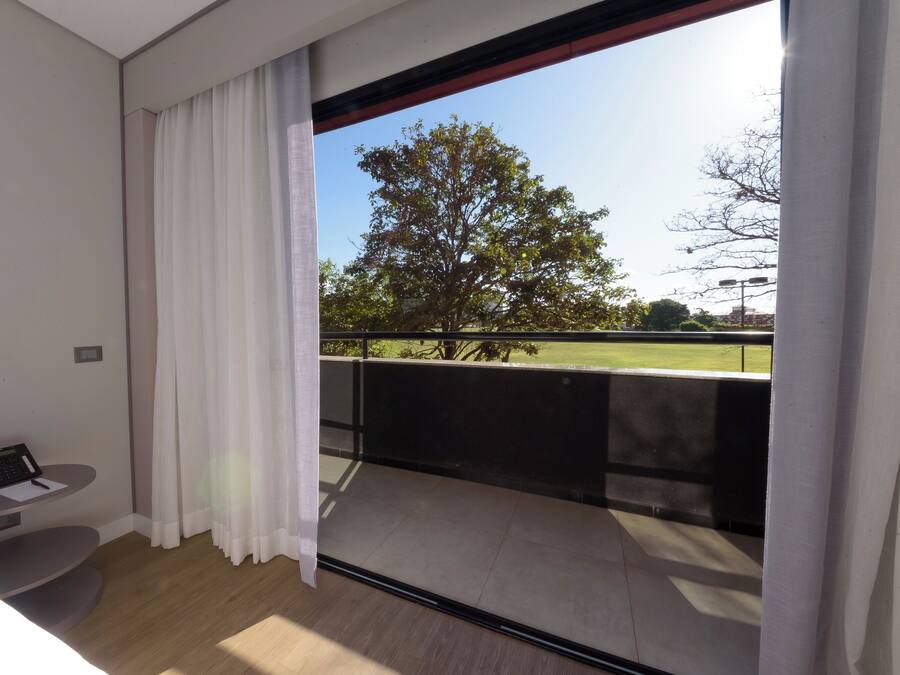 Ein modernes Hotelzimmer mit einem großen Fenster, einem kleinen Schreibtisch mit Telefon sowie Blick auf einen Park und Bäume.