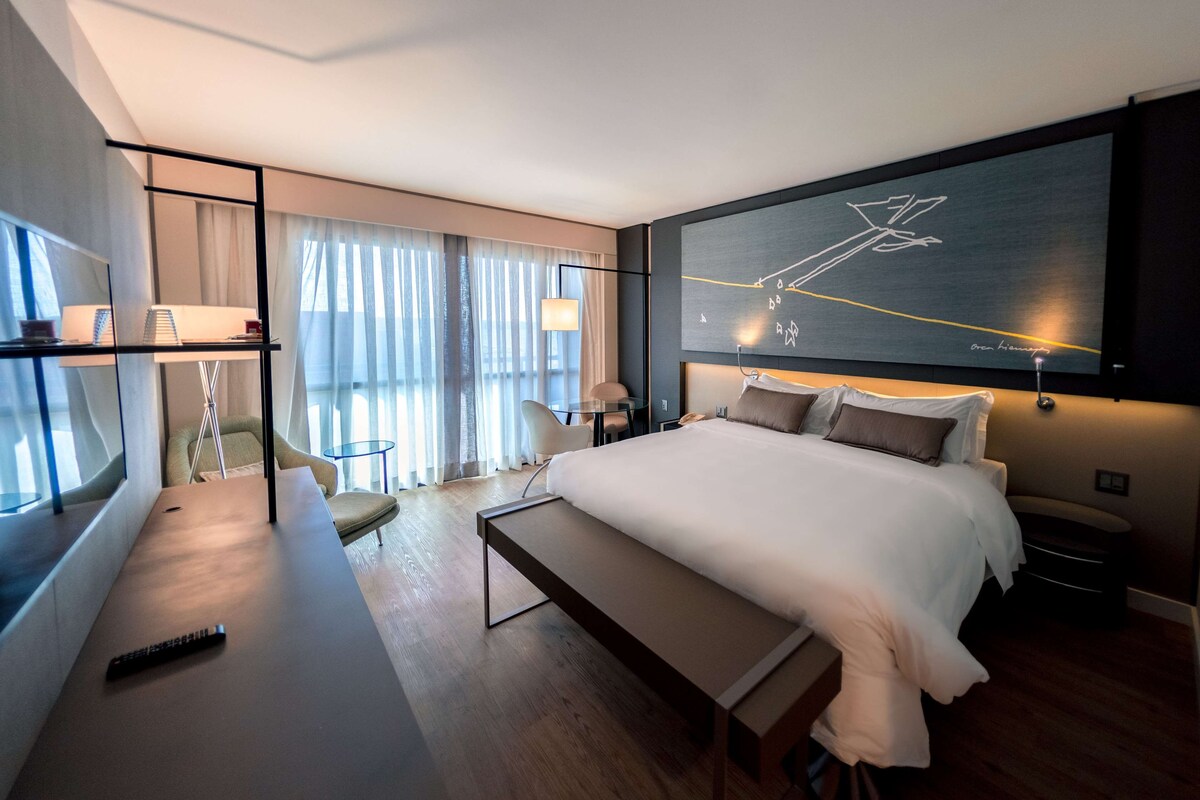 A modern hotel room with a large bed, a desk with a lamp, a chair, and a large window with curtains.
