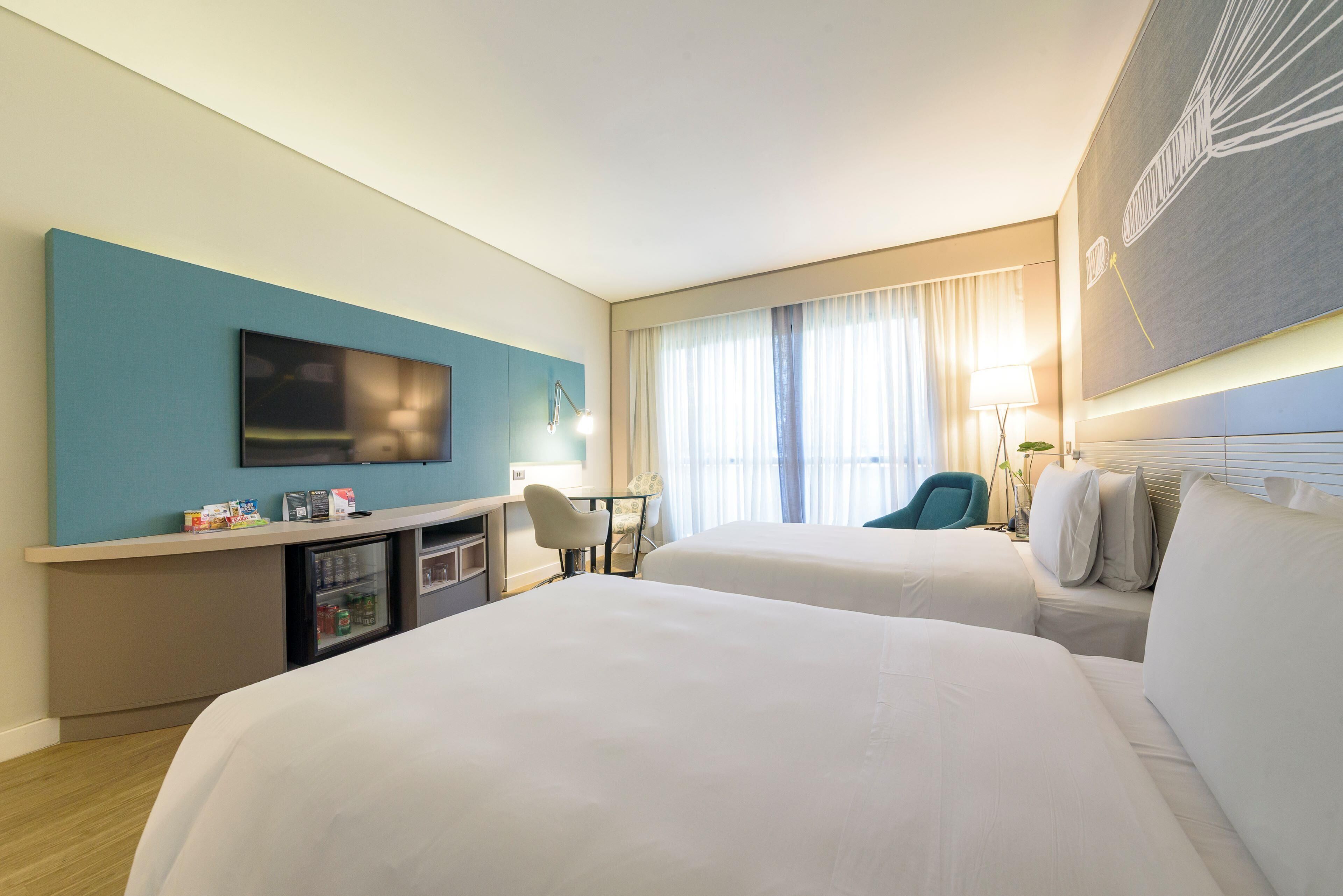Une chambre d’hôtel avec deux lits, un bureau avec une chaise, une télévision, un minibar et une grande fenêtre avec des rideaux.