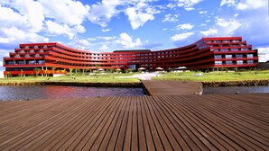 Un edificio moderno de color rojo con fachada curva, una terraza de madera en primer plano y una masa de agua al frente.