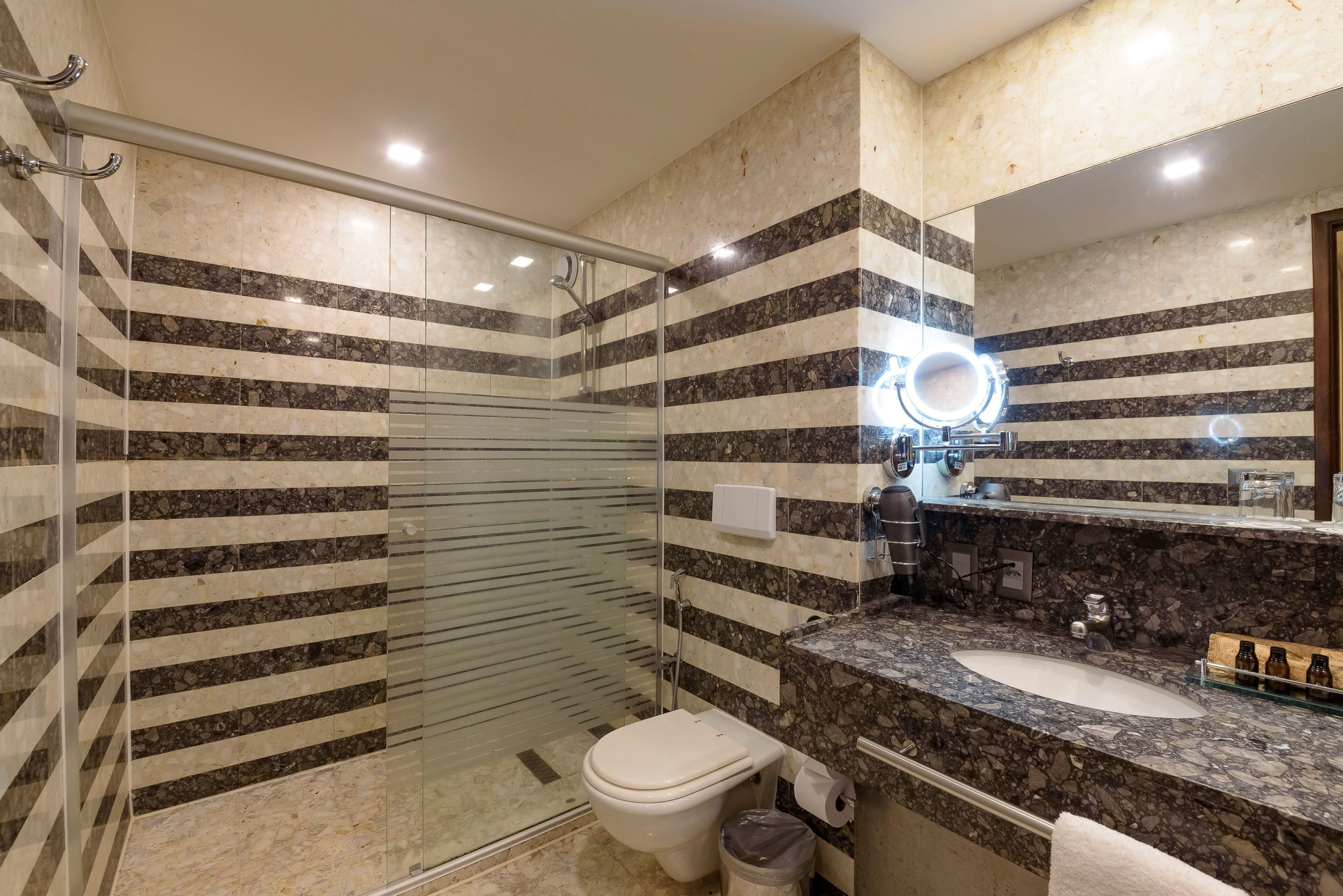 A modern bathroom with a glass-enclosed shower, a marble-tiled wall, a sink with a marble countertop, and a mirror with a circular light.