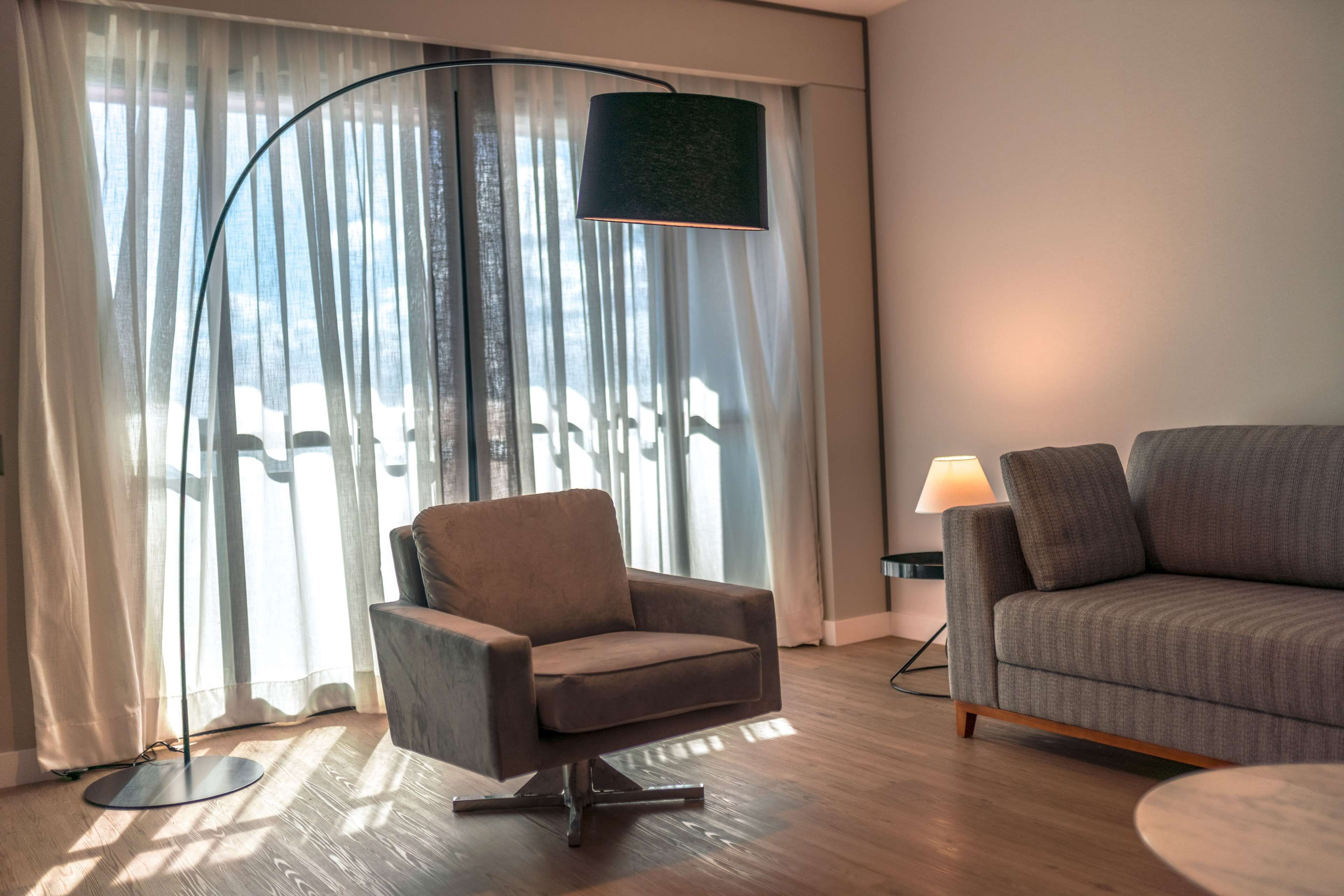 Una sala de estar moderna con un sillón gris, un sofá, una lámpara de pie y cortinas de gasa.