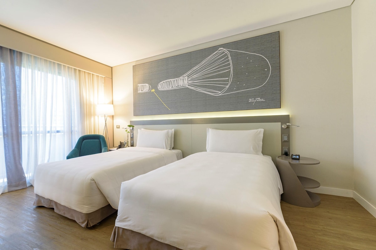 A hotel room with two beds, a desk, a chair, a lamp, and a wall art piece.