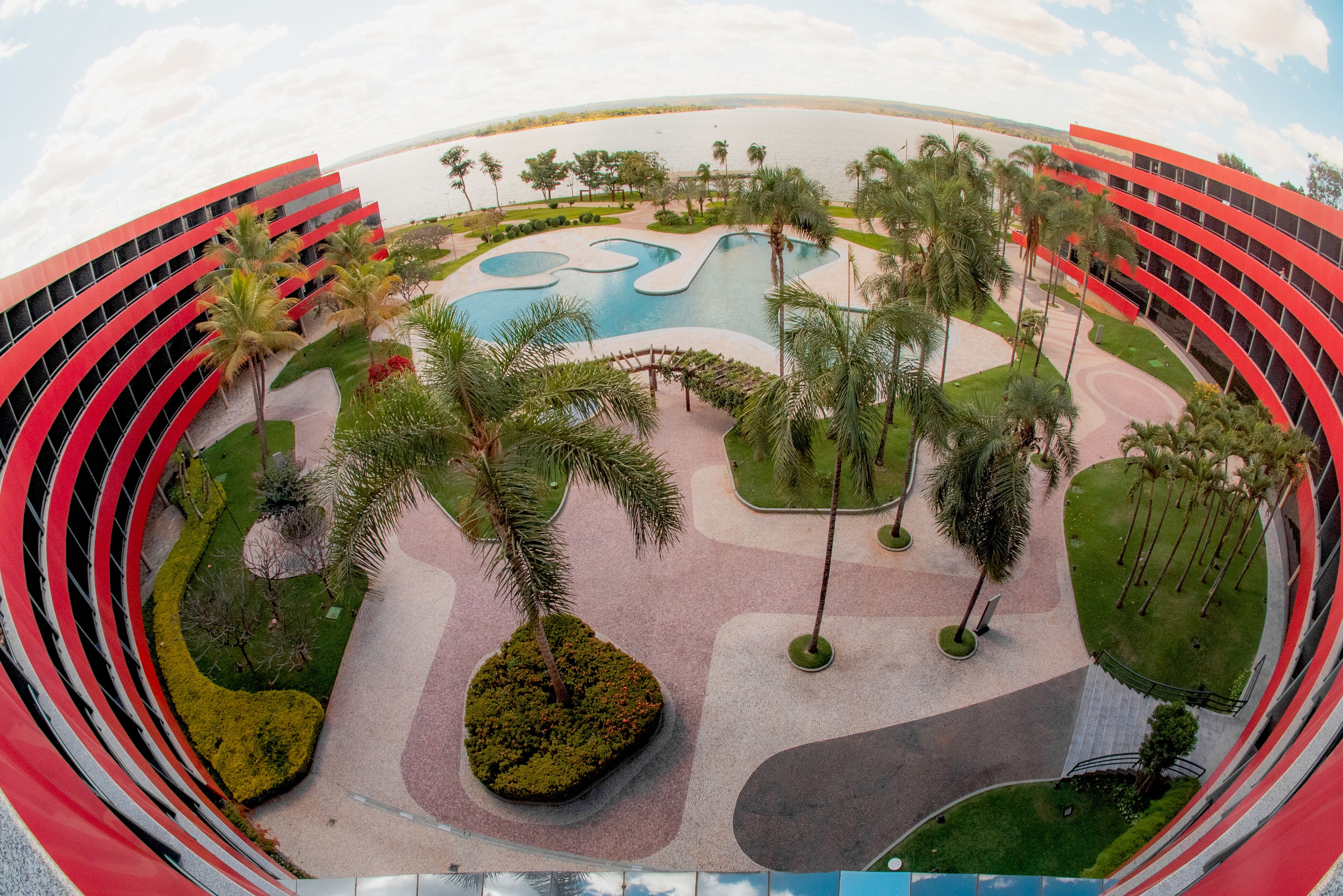 Un hôtel moderne doté d’une façade rouge incurvée, d’une piscine centrale et d’espaces paysagers environnants agrémentés de palmiers.