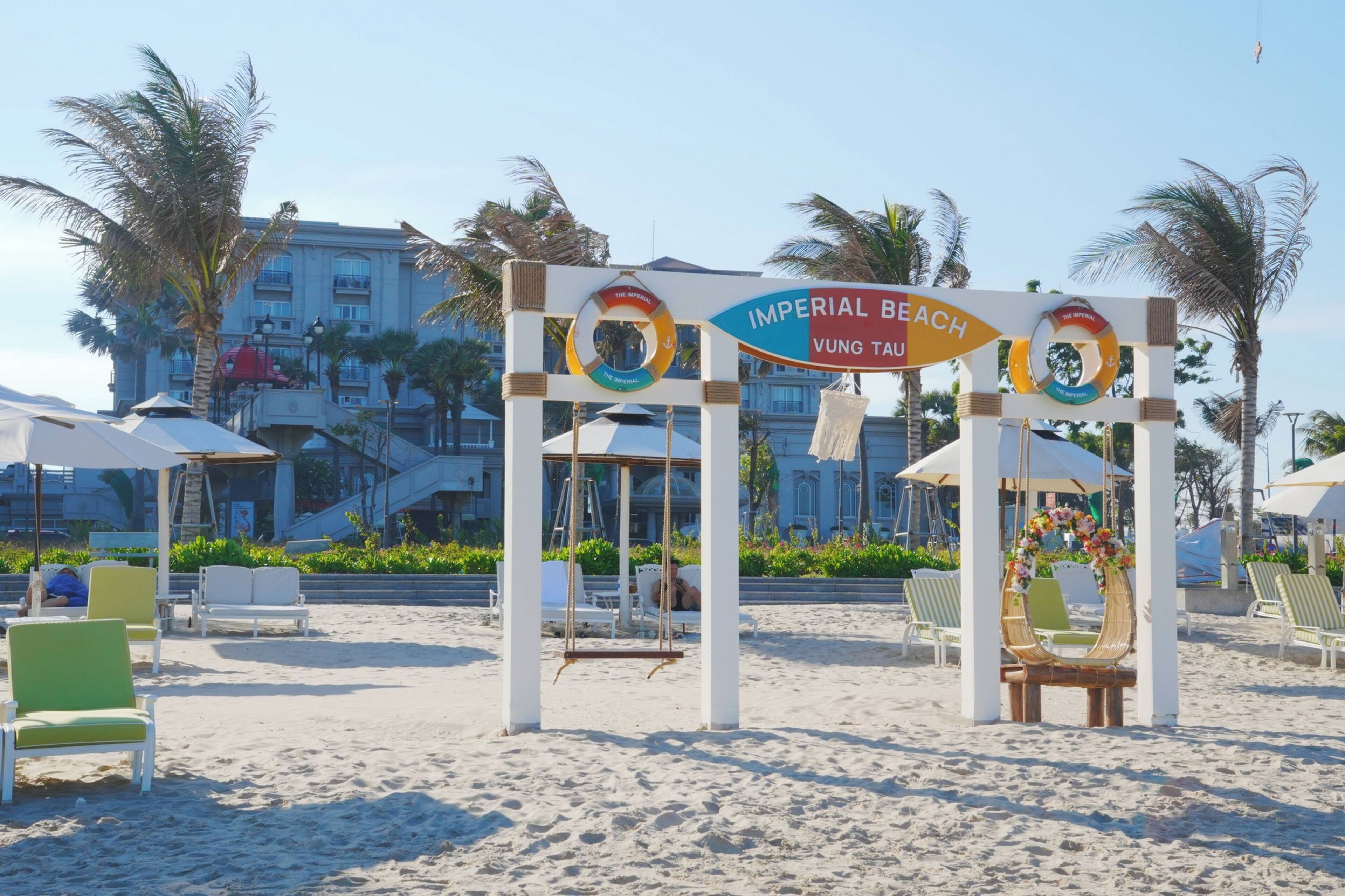 Zona de playa con un cartel que dice 'IMPERIAL BEACH VUNG TAU', palmeras y sillas de playa.