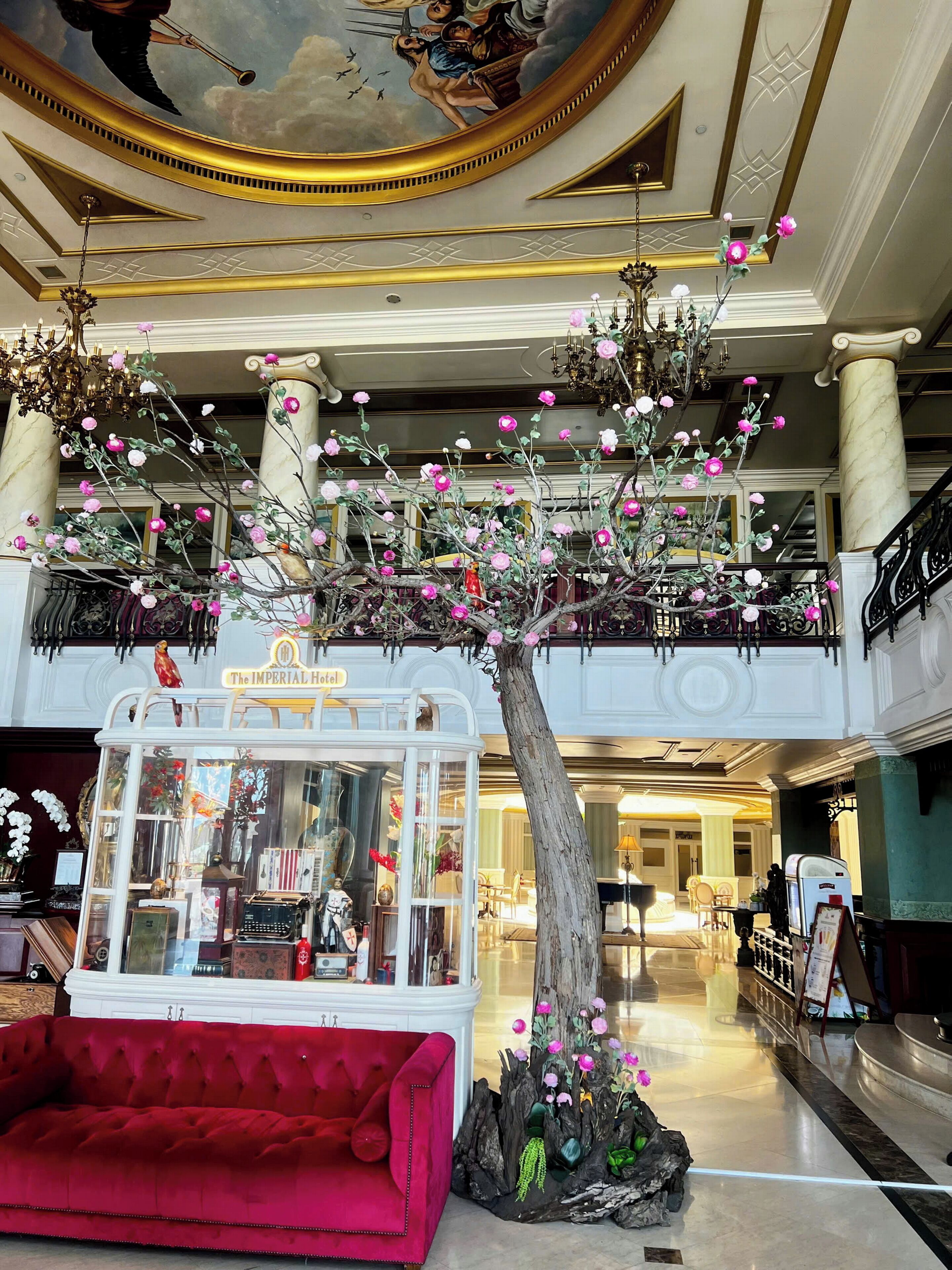 Um hall imponente com uma grande escultura de árvore, um sofá vermelho e um telefone público antigo com a inscrição 'The Imperial Hotel'.