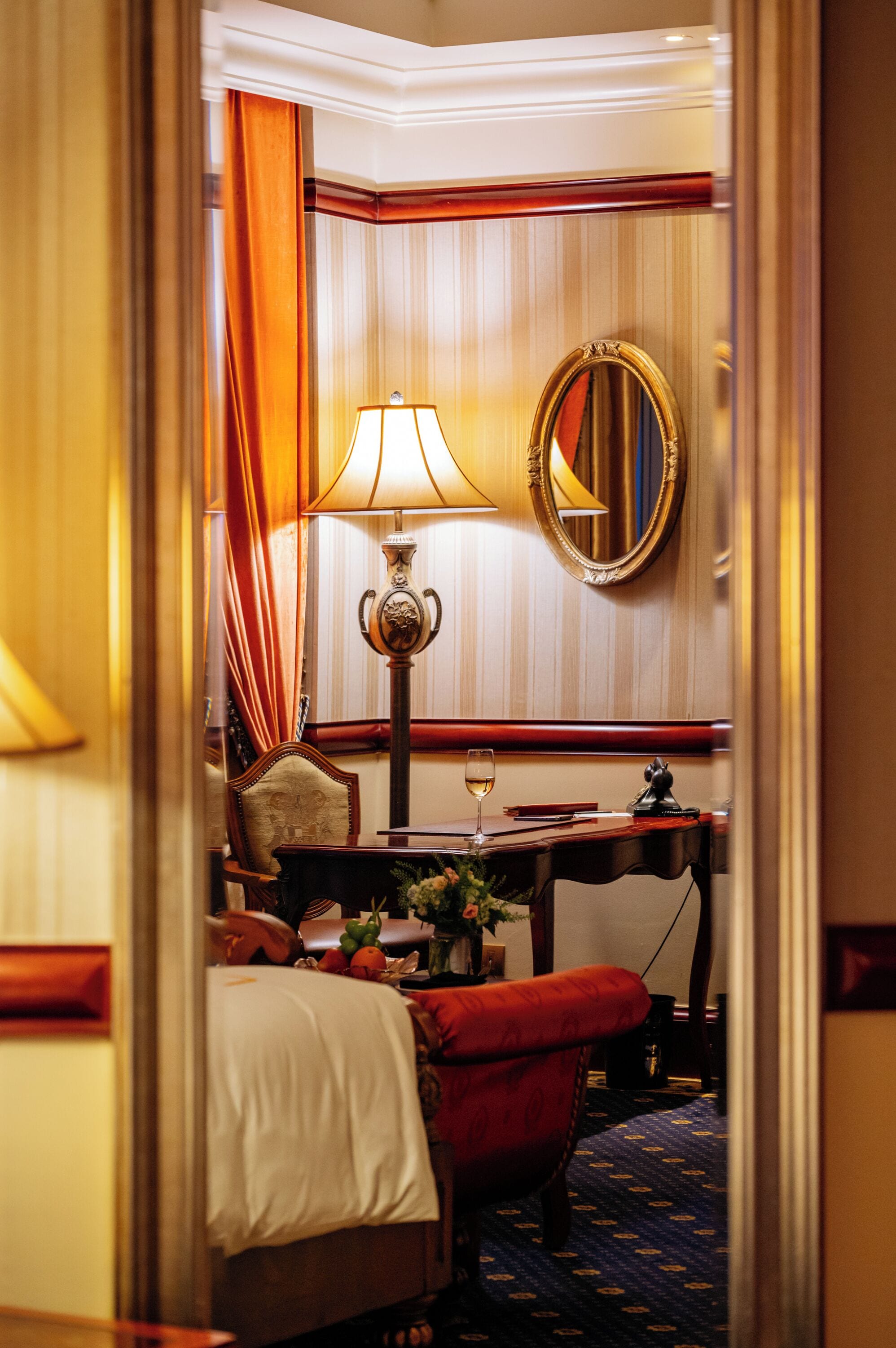 A hotel room with a bed, a red armchair, a table with a lamp, a mirror, and a vase with flowers.