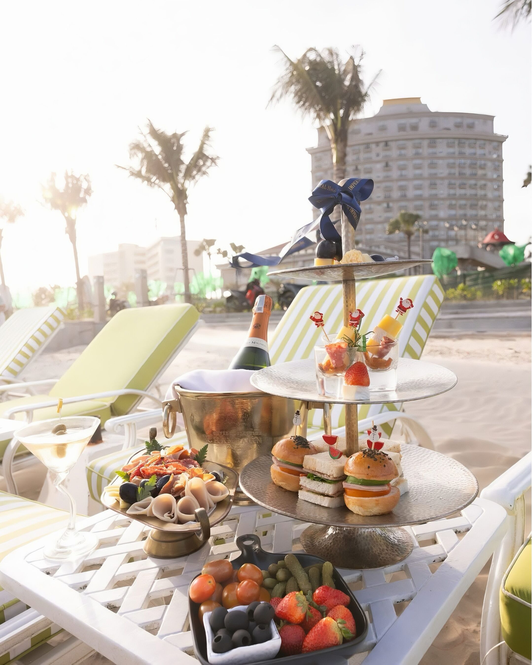 A beachside table with a variety of food and drinks, including sandwiches, fruits, and a bottle of champagne.