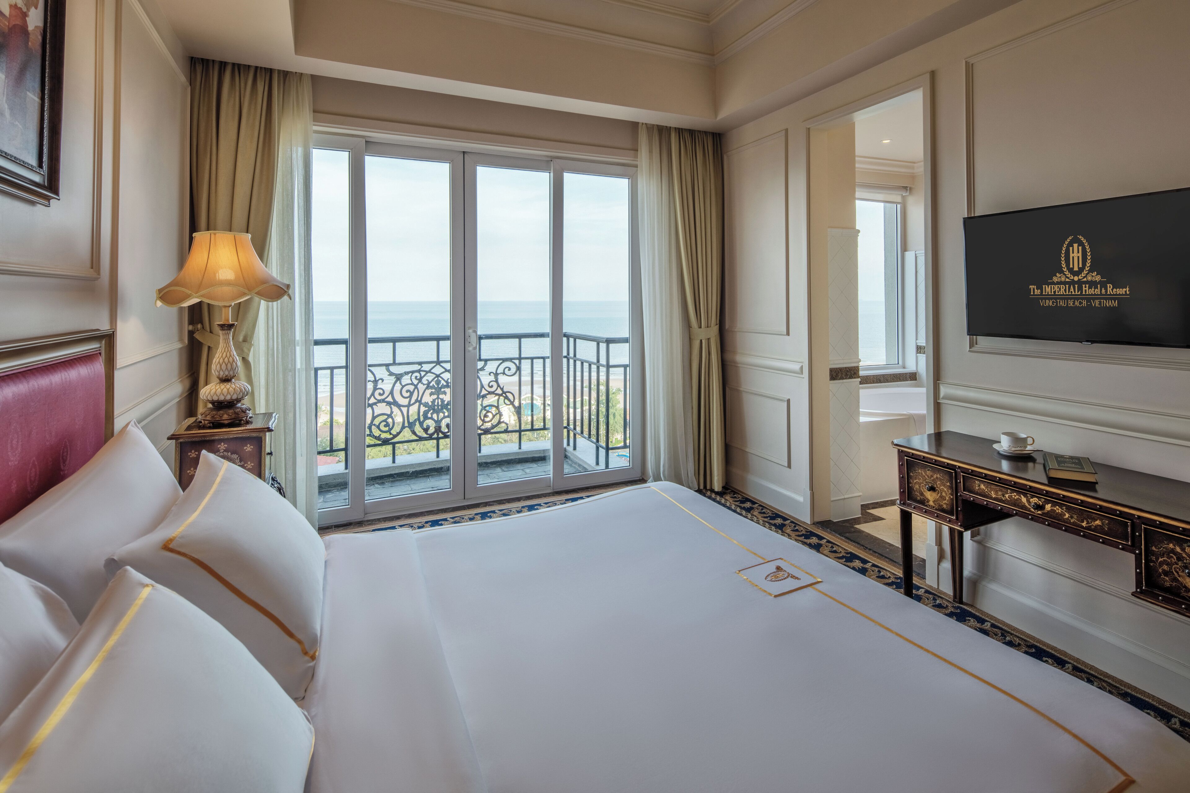 A hotel room with a large bed, a TV, a balcony with a view, and a desk with a cup and book.