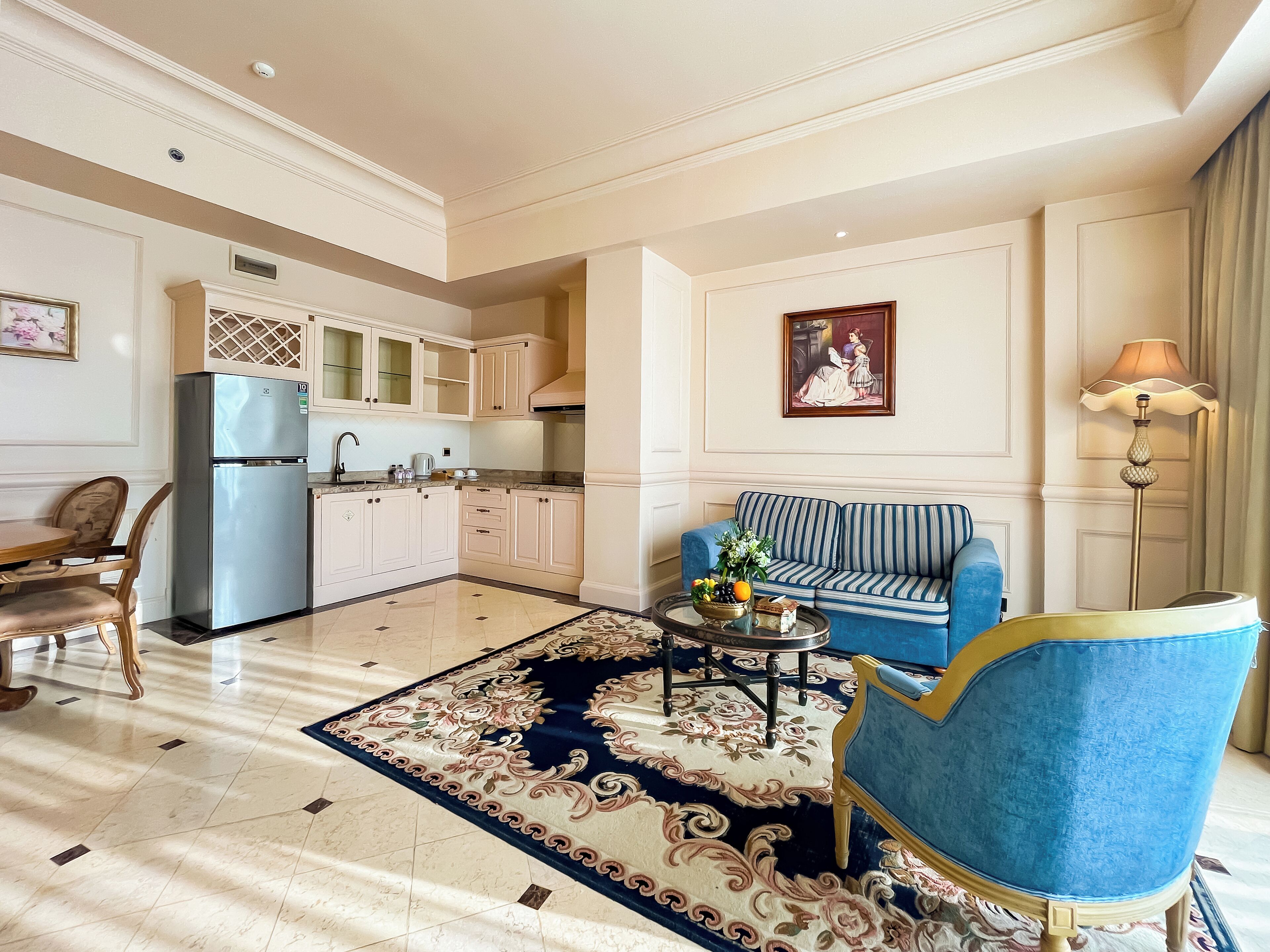 Amplia sala de estar con un sofá azul, una mesa de centro con frutas y un florero, una mesa de comedor con sillas y una cocina al fondo.