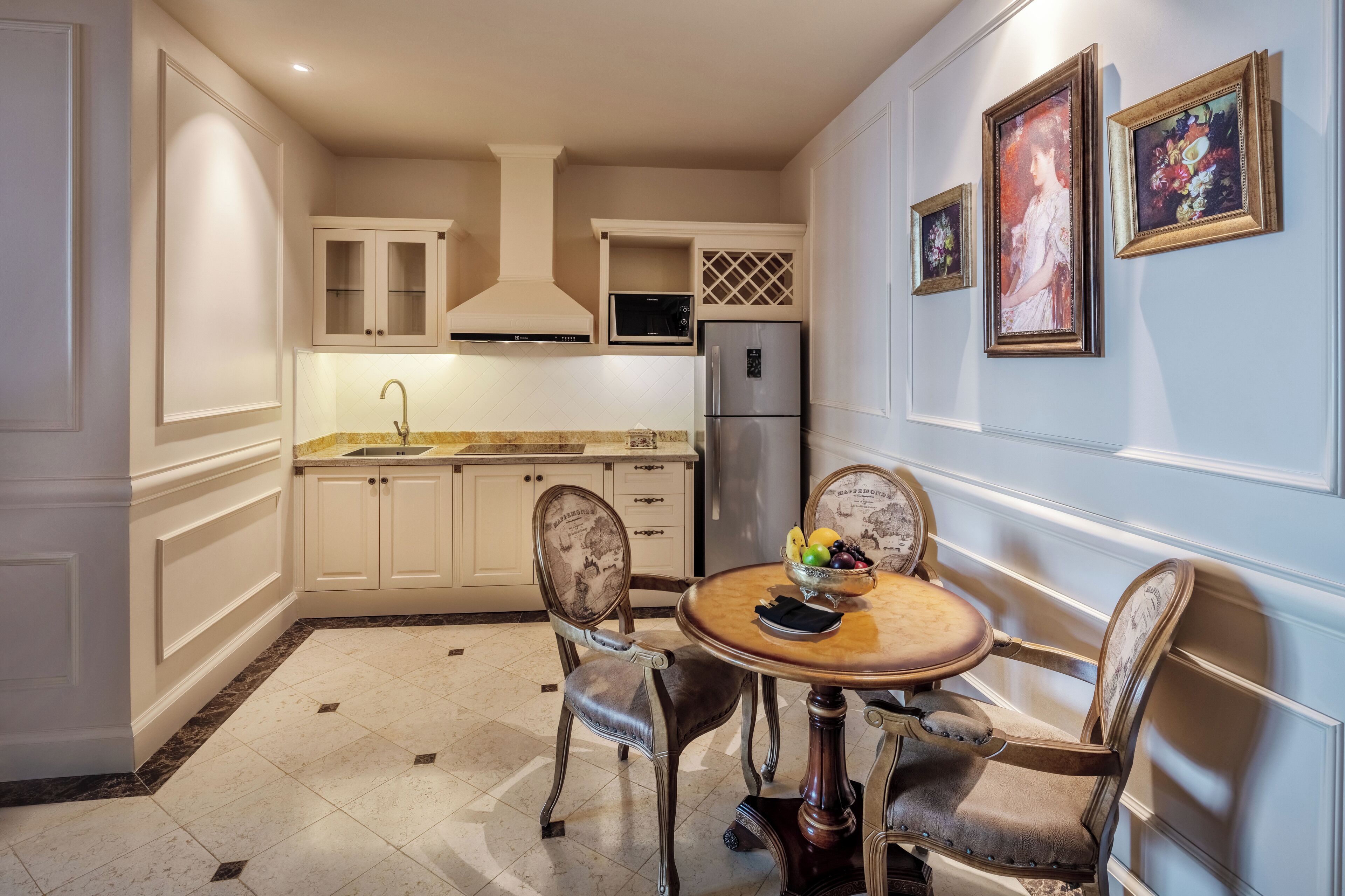 A compact kitchen with white cabinetry, a stainless steel refrigerator, a microwave, and a sink. There is a dining table with chairs, and a bowl of fruit on the table.