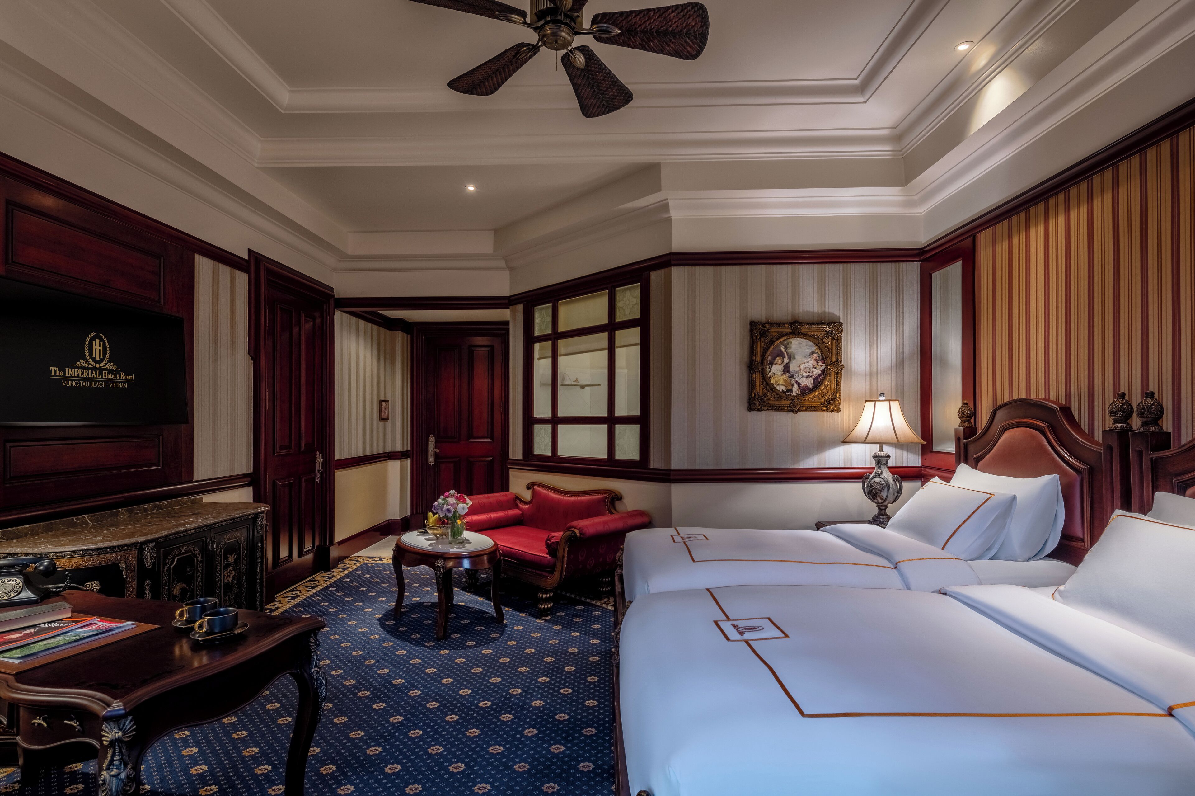 A hotel room with a large bed, a desk with a telephone, a red armchair, a television, and a ceiling fan.