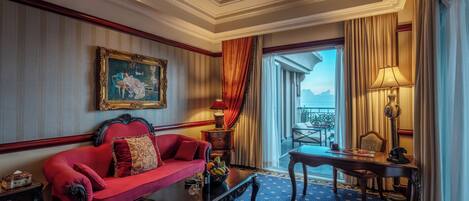 A hotel room with a red sofa, a wooden coffee table, a painting on the wall, a balcony with a view, and a ceiling fan.