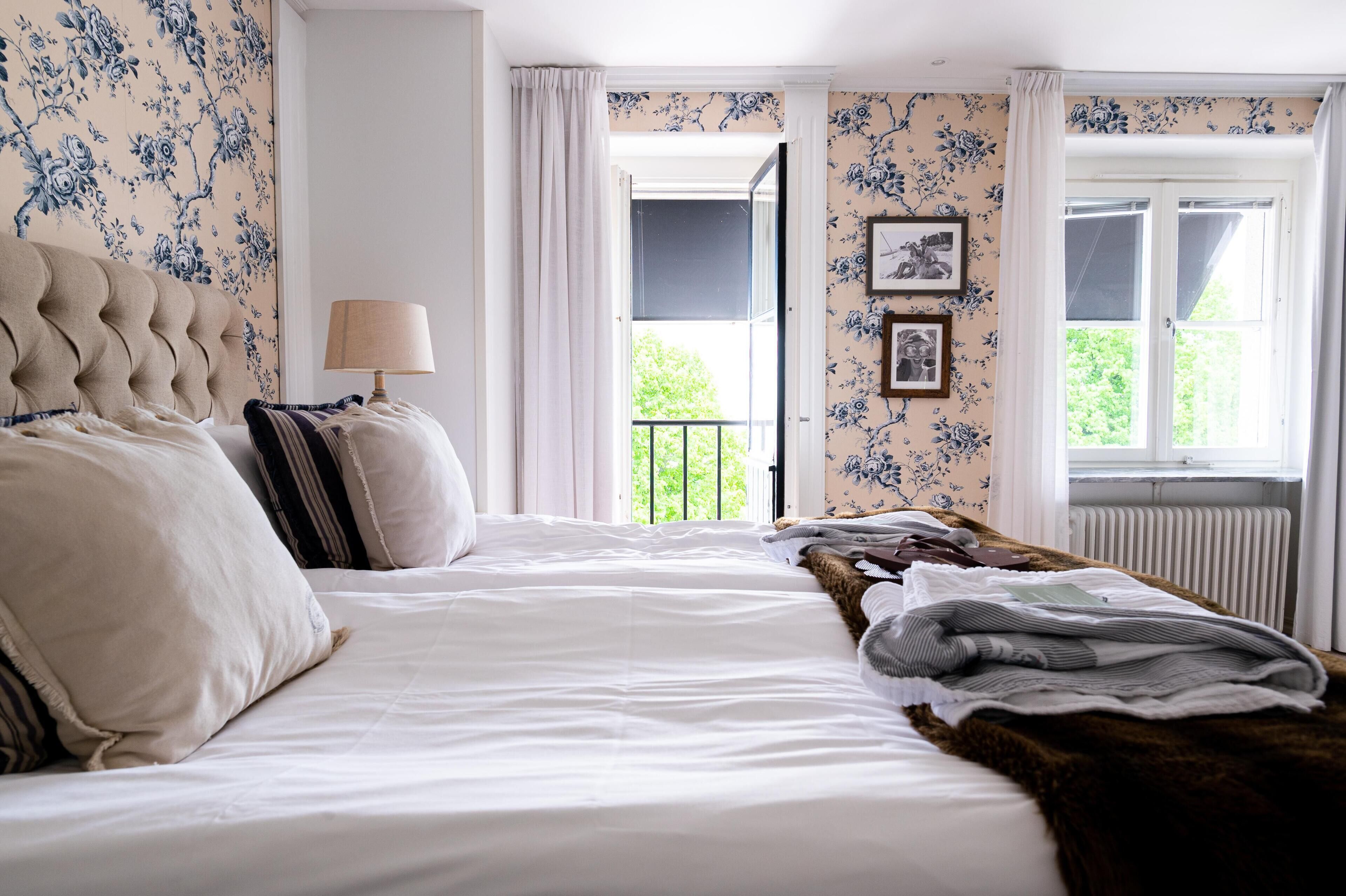 A bedroom with a large bed, a bedside table with a lamp, floral wallpaper, and a view of the outdoors through a window.