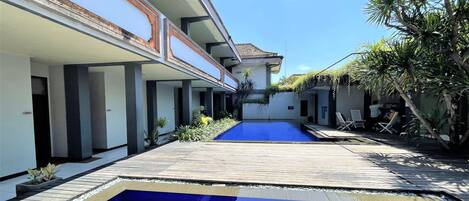 Zona de piscina con terraza de madera, piscina azul cristalina y un edificio con múltiples balcones.