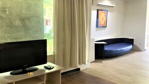 A living room with a flat-screen TV on a stand, a blue sofa, and a window with curtains. - The Yani Hotel Bali (Denpasar)