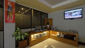 A modern hotel lobby with a buffet area, a large window with decorative tiles, a television mounted on the wall, and a potted plant. - The Yani Hotel Bali (Denpasar)