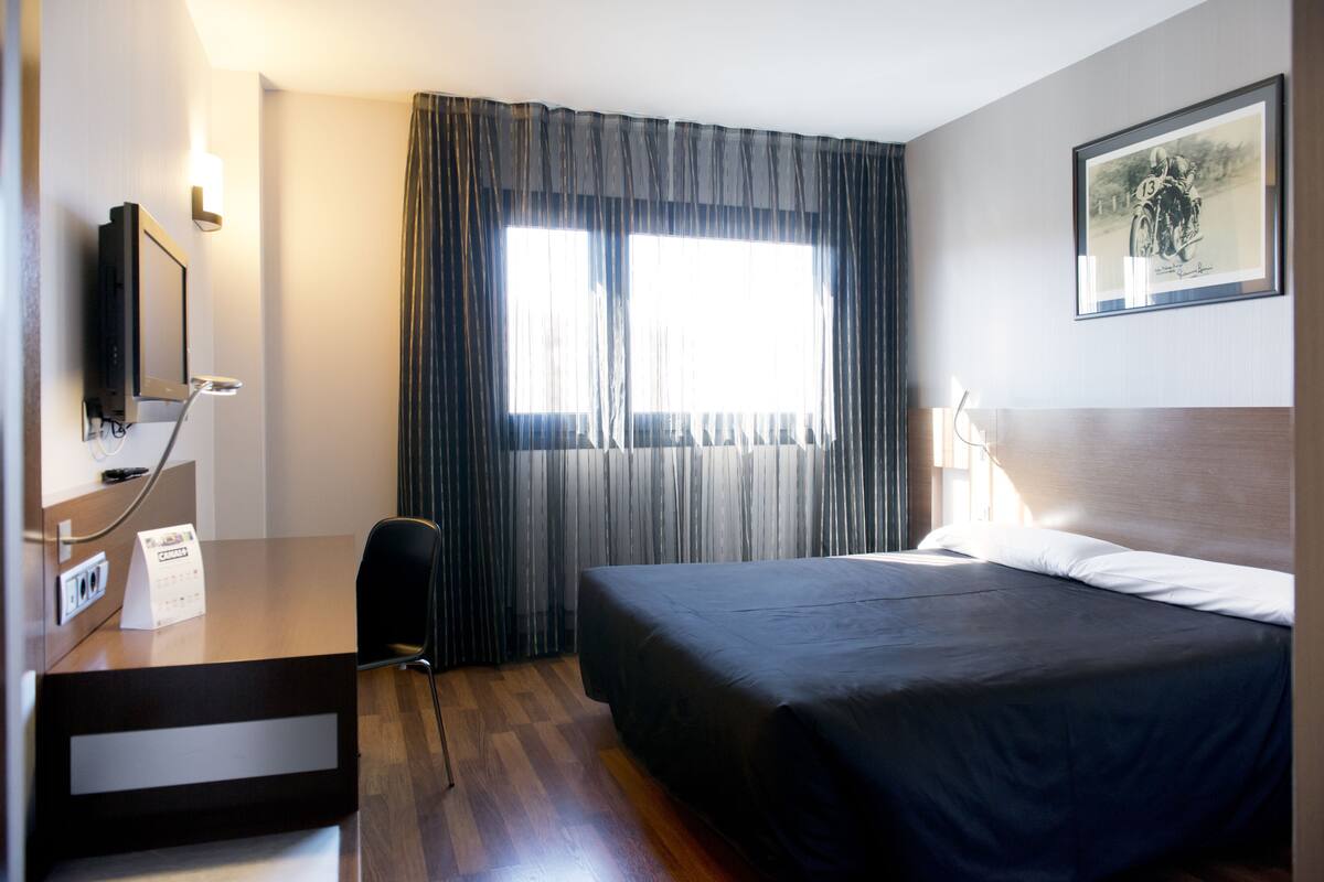 Une chambre d’hôtel avec un lit, un bureau avec un ordinateur, une chaise et une fenêtre avec des rideaux.