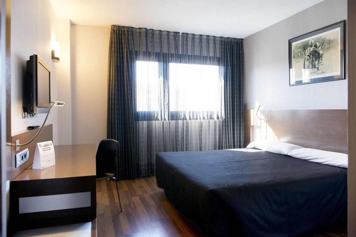 Ein Hotelzimmer mit einem Bett, einem Schreibtisch mit Computer, einem Sessel, einem Fenster mit Vorhängen und einem Bild im Bilderrahmen an der Wand.