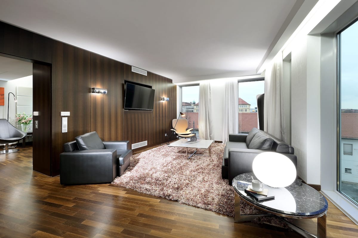 A modern hotel room with a wooden accent wall, a flat-screen TV, a sofa, a coffee table with a round mirror, and a large window offering a city view.