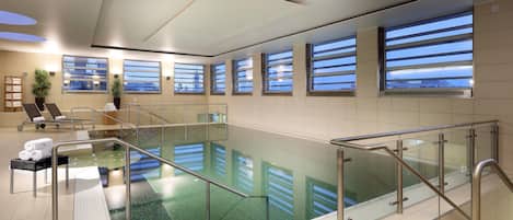 Una piscina interior moderna con barandillas de vidrio transparente, un techo iluminado por el sol y amplias ventanas con vistas al cielo.