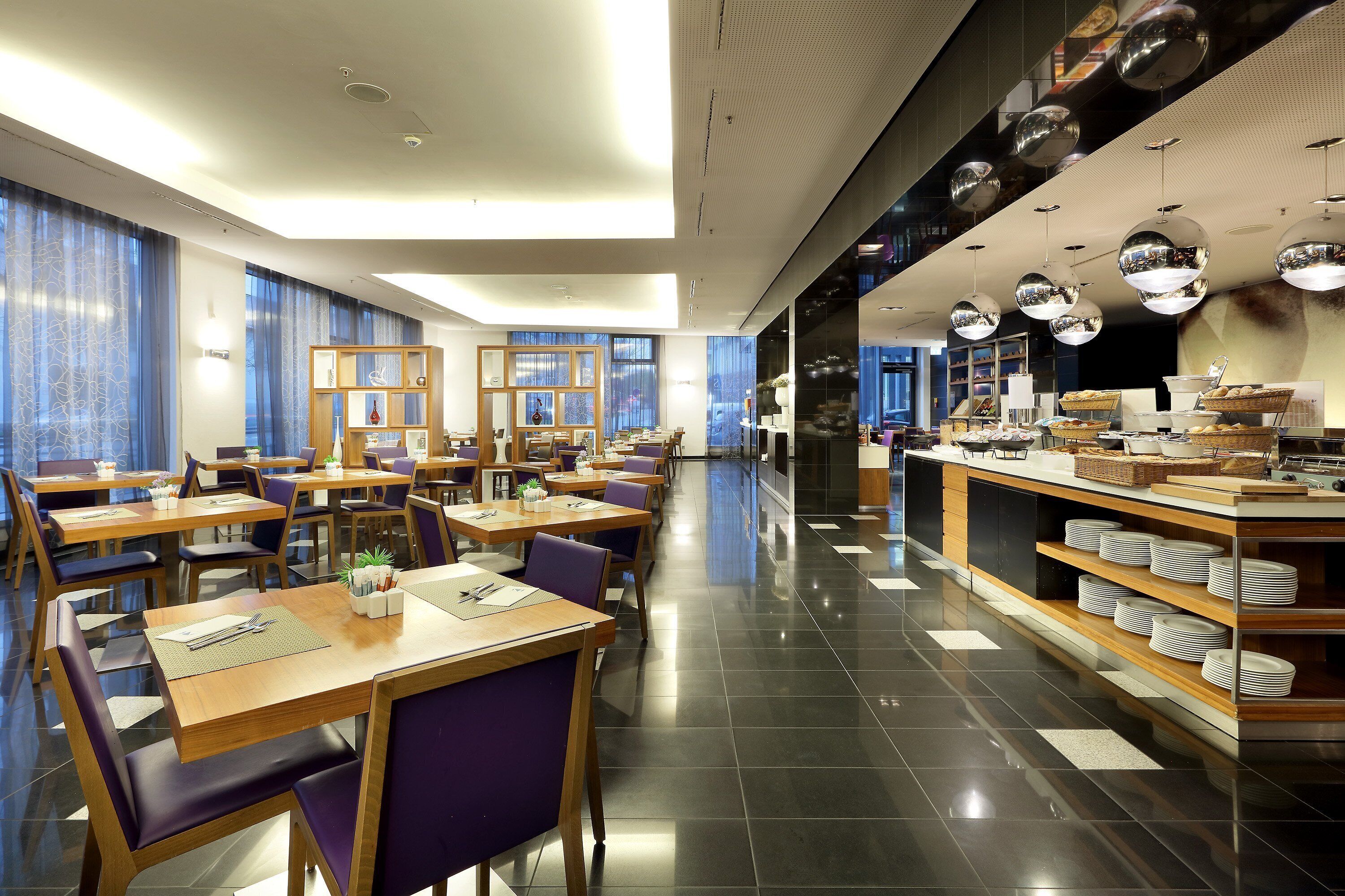 Un restaurant modern amb taules de fusta i cadires morades, una zona de buffet amb plats i cistos, i grans finestres amb cortines blaves.