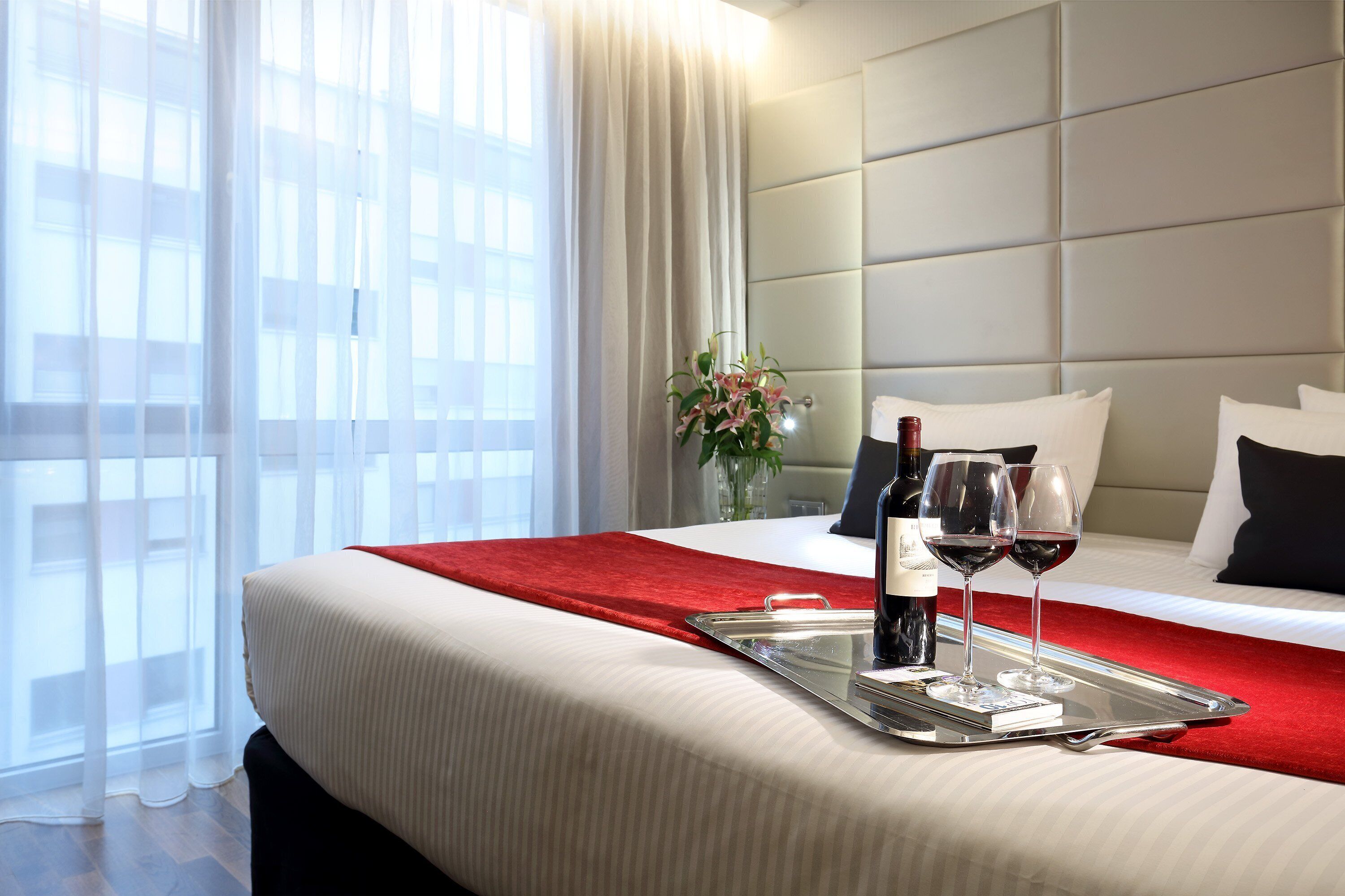 A hotel room with a bed, a tray with wine glasses and a bottle, and a vase with flowers.
