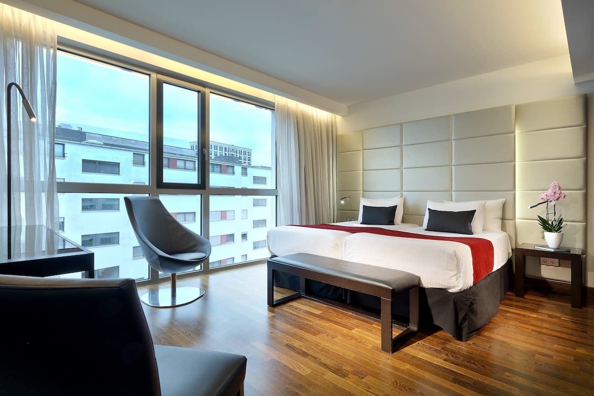 A modern hotel room with a large bed, a desk, a chair, and a view of the city through large windows.