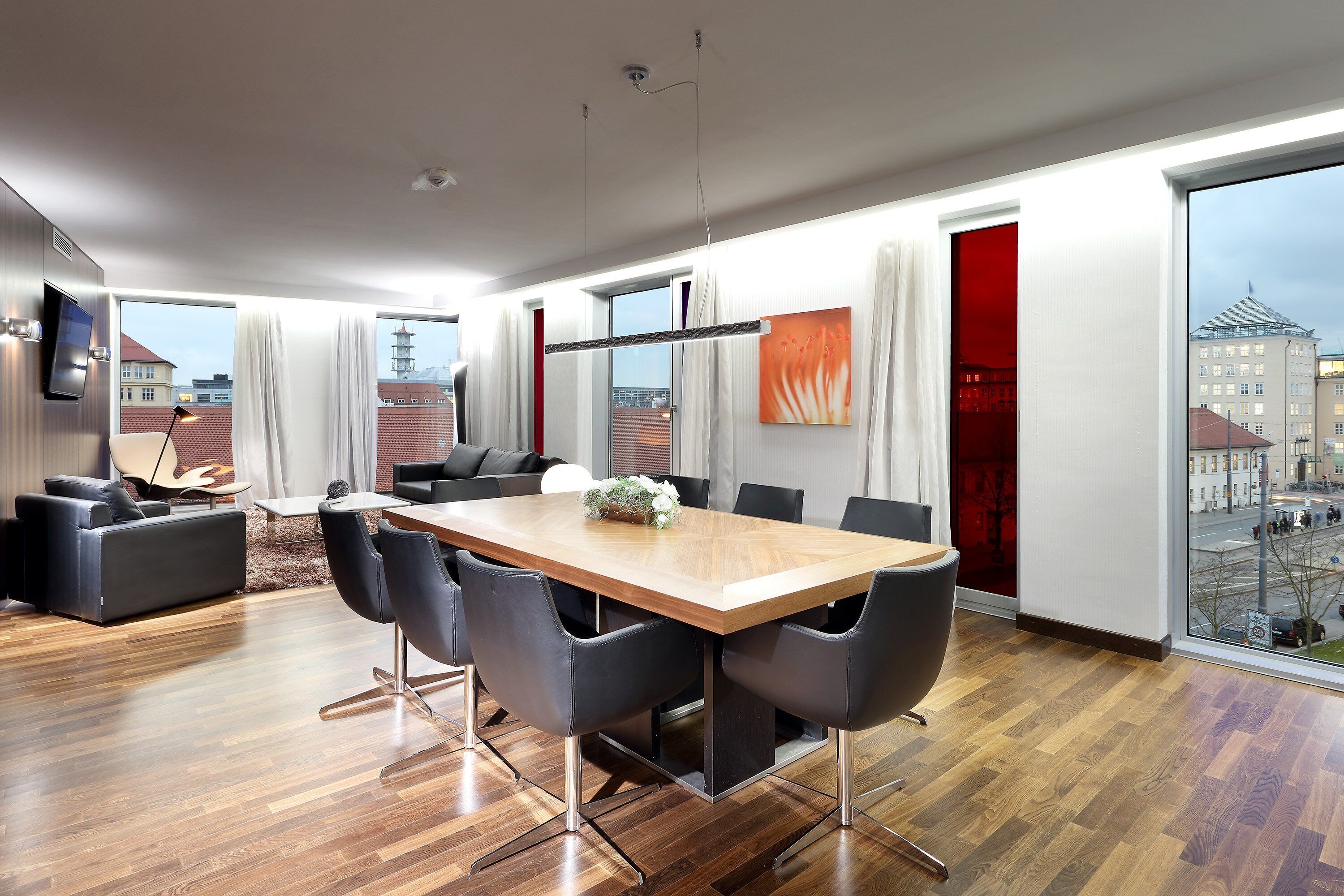 Una habitación de hotel moderna con una mesa de comedor de madera, sillas negras, un sofá y un televisor montado en la pared.