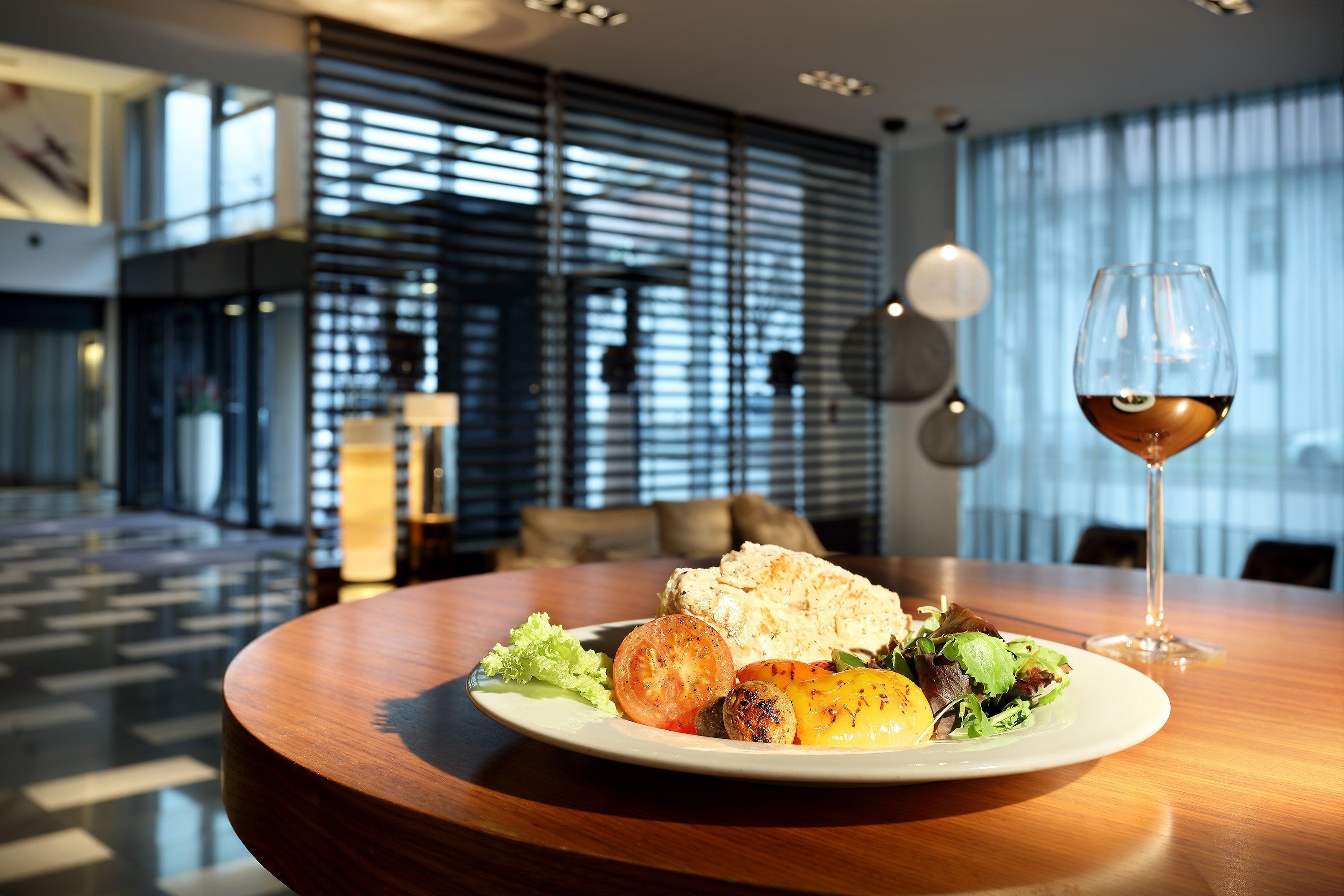 A dining table with a plate of food, a glass of red wine, and a modern interior with large windows and a patterned floor.