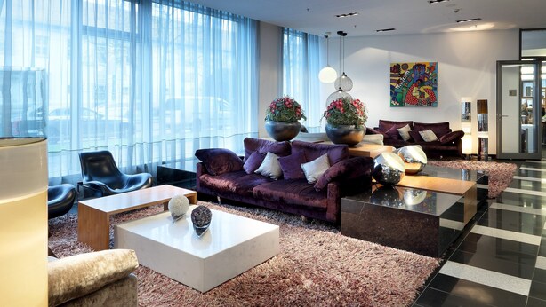 Una sala de estar moderna con un sofá morado, sillas negras, una mesa de centro blanca y un gran ventanal con cortinas translúcidas.